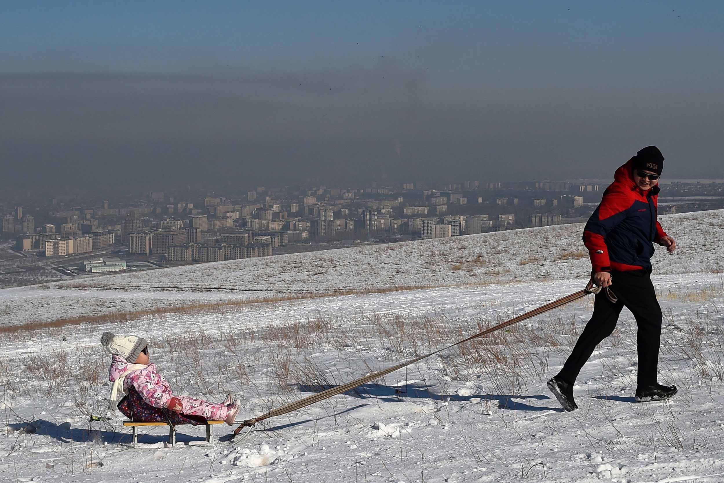A man pulls a child on a sled on a hilltop as smog covers the Kyrgyz capital of Bishkek on January 29, 2020. © VYACHESLAV OSELEDKO/AFP via Getty Images