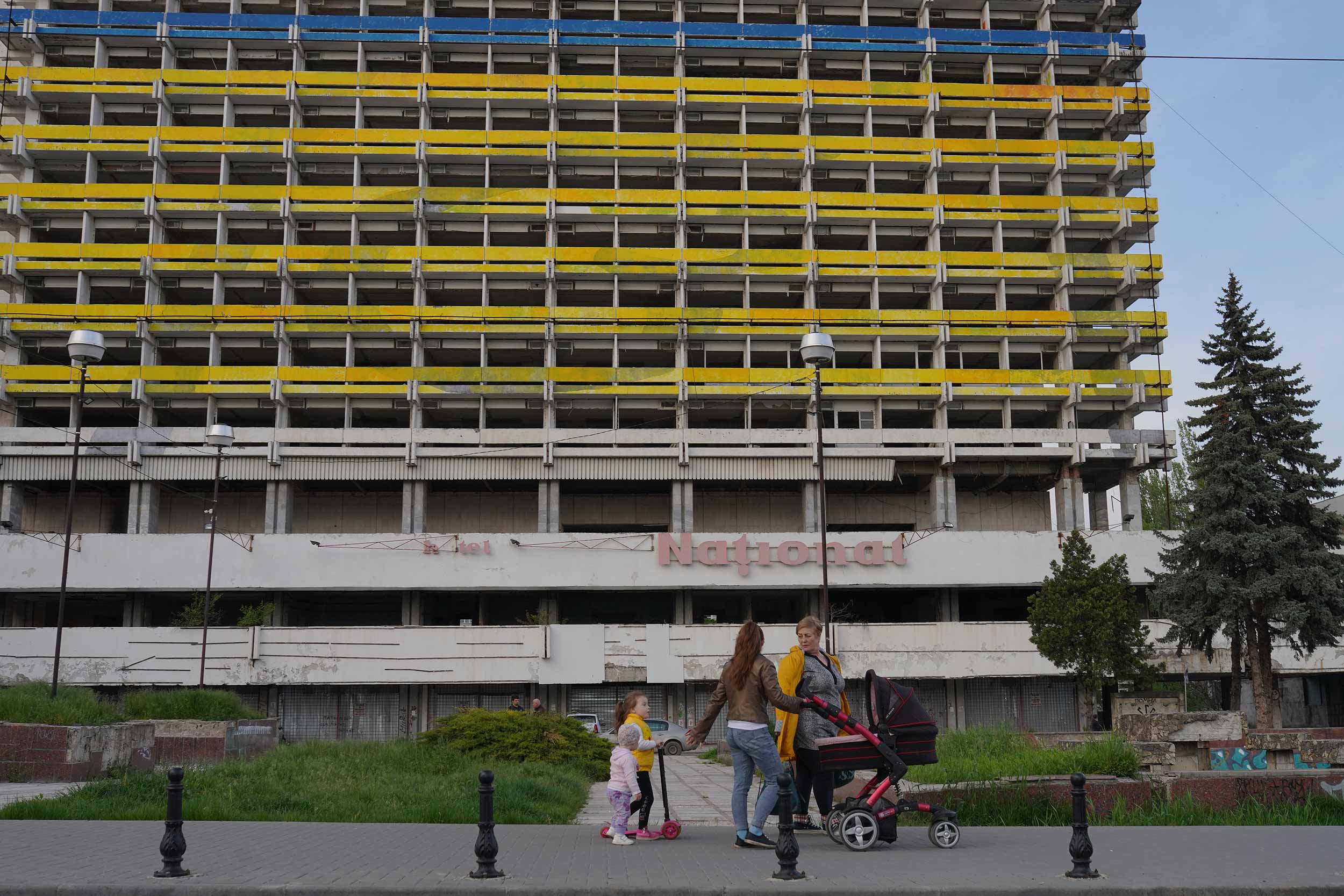 A family walks past the Soviet-era hotel National, now abandoned and painted in the colours of the Ukrainian flag, on May 3, 2022 in Chisinau, Moldova. More than two months into the Russian invasion of Ukraine, fears of the conflict spilling over into Moldova, a country of 2.5 million inhabitants, has increased after two attacks on April 23 and 24 in the separatist region of Transnistria. © Andreea Campeanu/Getty Images