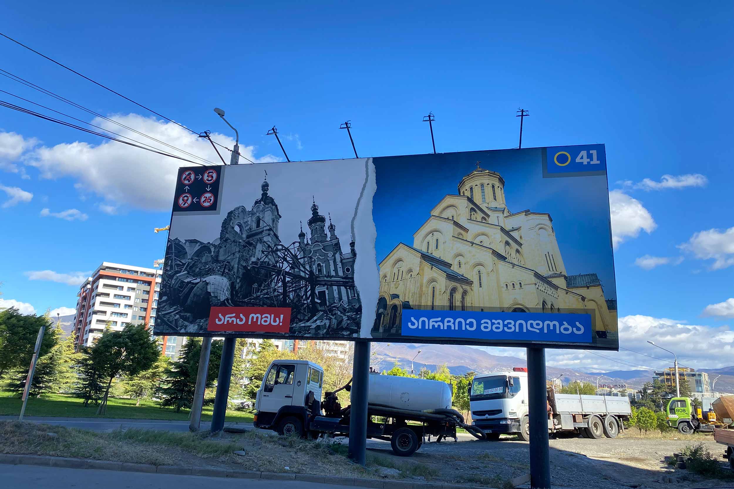 One of a series of Georgian Dream election banners depicting images of war-torn Ukraine with colourful photos of a prosperous Georgia. On the left is an Orthodox church destroyed by Russian shelling in Ukraine – and the ballot paper numbers of the four opposition parties - while on the right is the Holy Trinity Church in Georgia, built with billionaire Bidzina Ivanishvili's financial support. The left side of the banner reads "NO WAR," and the right says, "Elect/Choose Peace." © IWPR